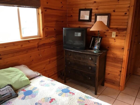 A wooden cabin room with a bed, a dresser, a television, and framed pictures on the wall. - Cute Lakefront Cottage on Lake Michigan. (Mears)