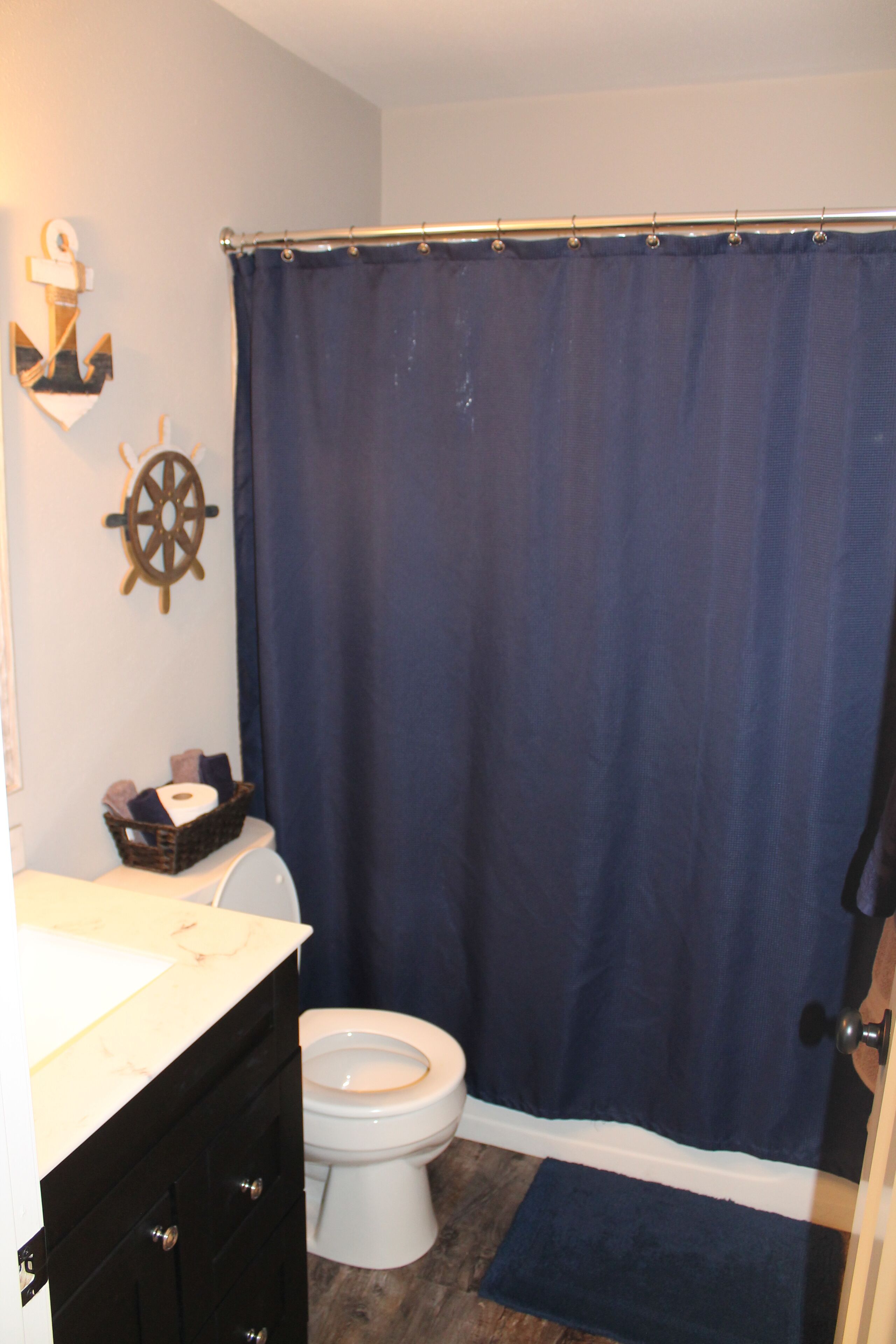 A bathroom with a white toilet, a dark blue shower curtain, a wooden ship wheel decoration, and a basket with towels on the counter.