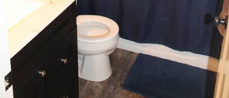 A bathroom with a white toilet, a dark blue shower curtain, a wooden ship wheel decoration, and a basket with towels on the counter.