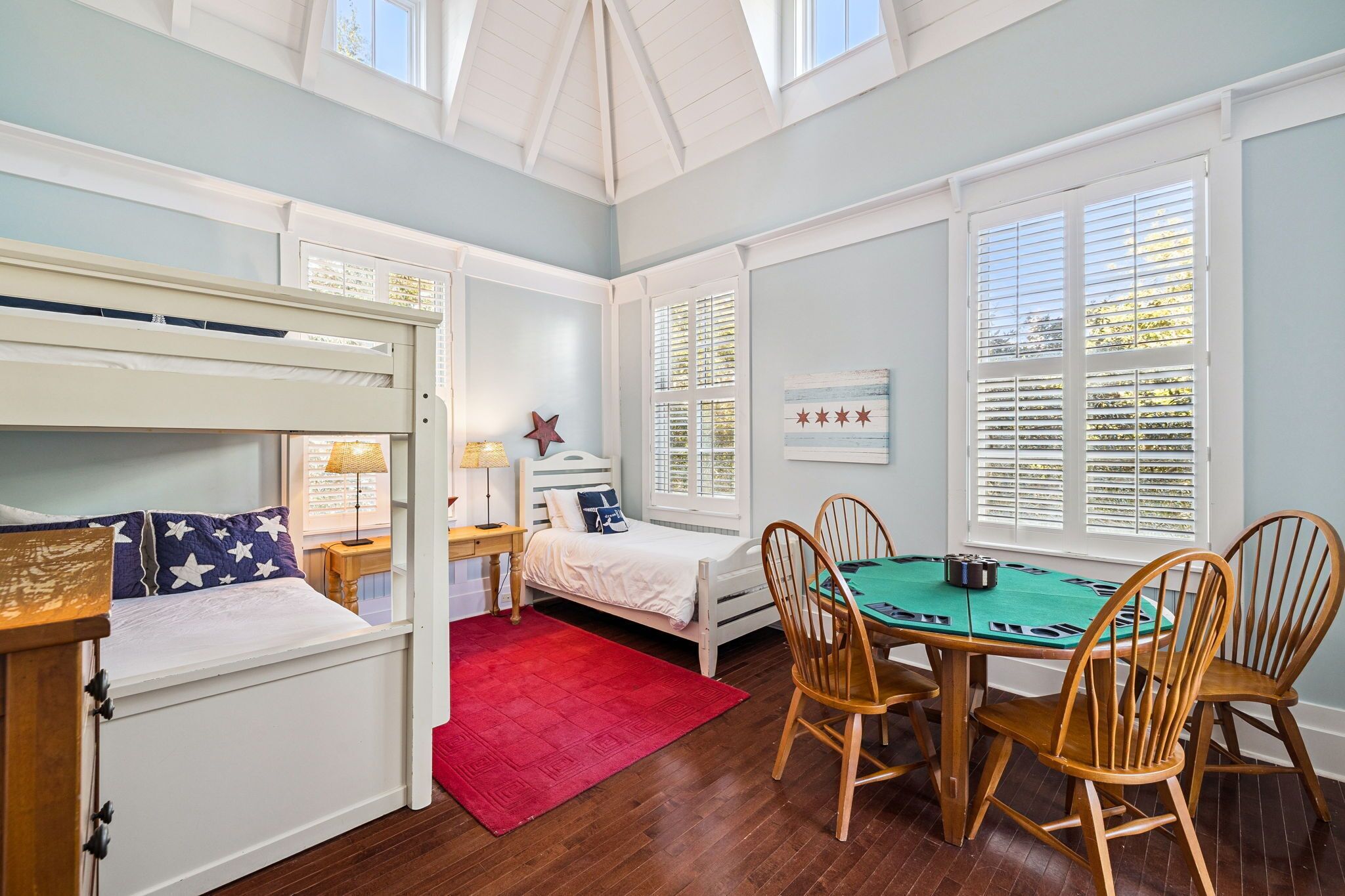 A room with a bunk bed, a desk with a lamp, a small table with chairs, and a window with blinds.