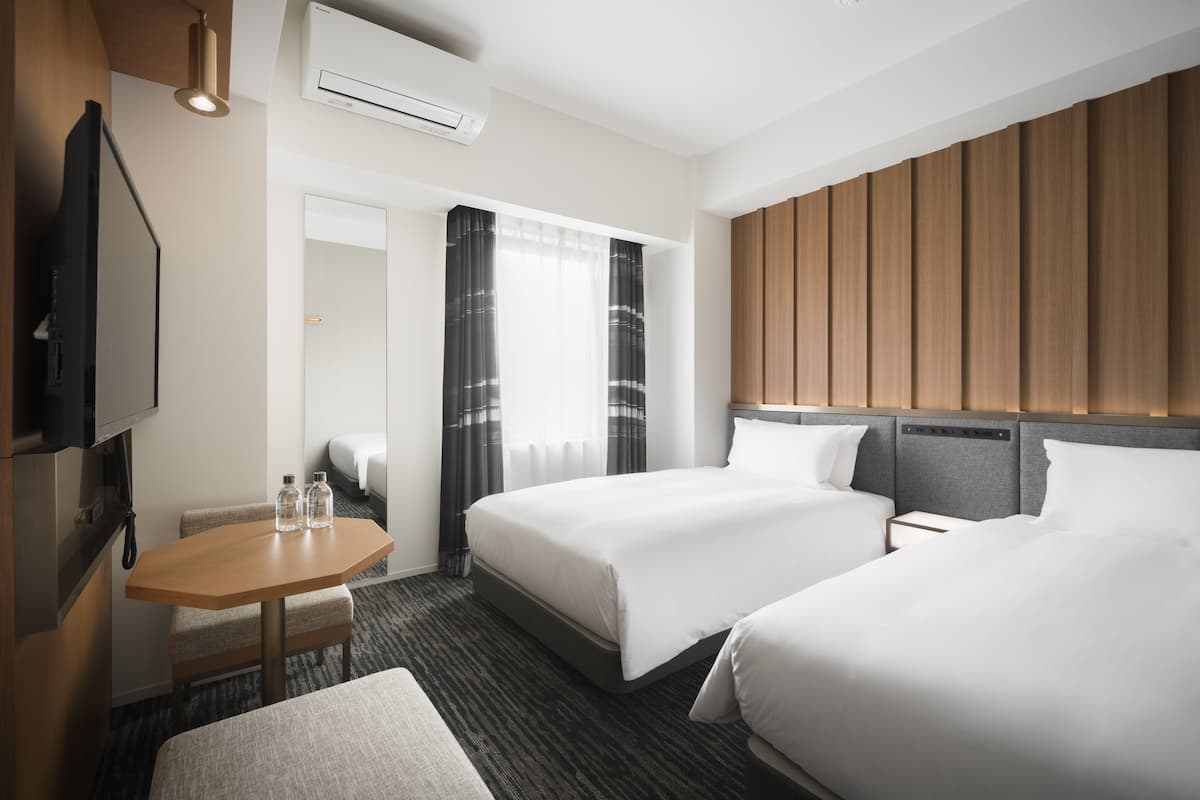 A hotel room with two beds, a TV, a desk, and a window with curtains.