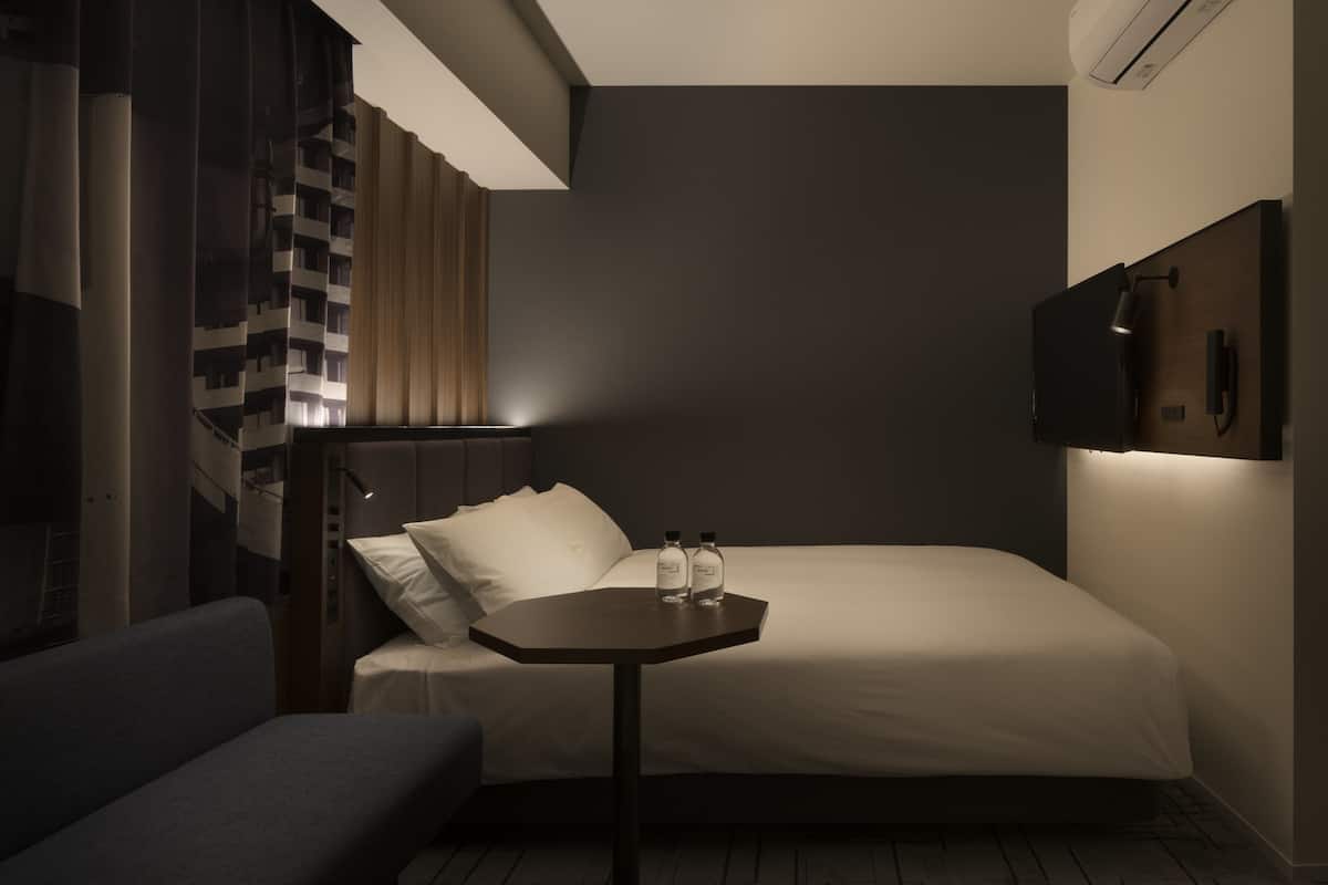 A modern hotel room with a bed, a small table with two bottles, a sofa, and a TV mounted on the wall.
