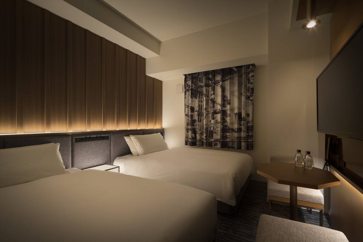 A hotel room with two beds, a small table with two bottles, a TV, and a patterned wall.