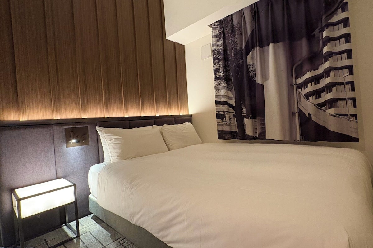 A modern hotel room with a large bed, a nightstand, a wall-mounted light, and a large window with a view of a building.