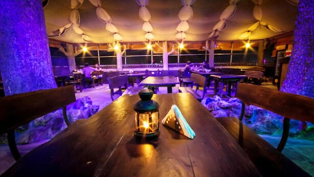 A dimly lit restaurant with wooden tables and benches, a lantern on one table, and a view of the outside through large windows. - Hotel Ruby Arena (Thiruvananthapuram)