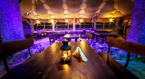 Un restaurante tenuemente iluminado con mesas y bancos de madera, un farol sobre una mesa, y vistas del exterior a través de grandes ventanales.