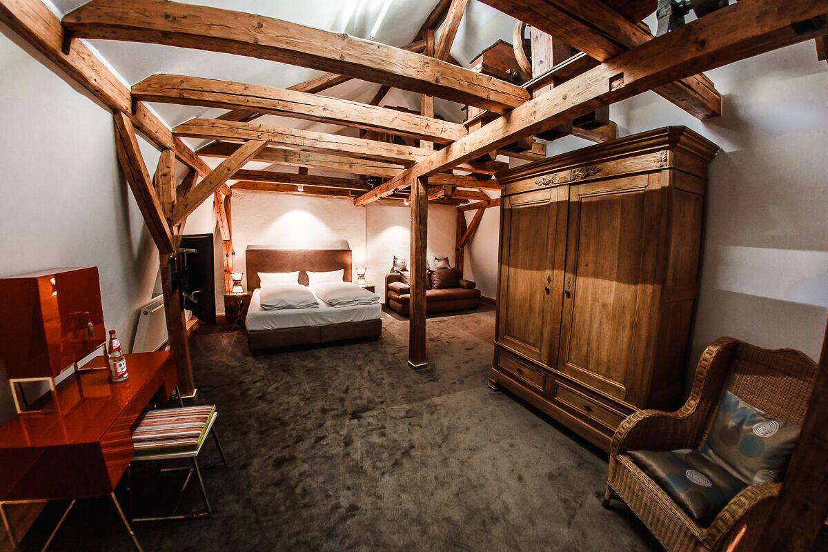 A bedroom with a sloped ceiling, wooden beams, a bed, a wardrobe, a chair, and a desk with a lamp.