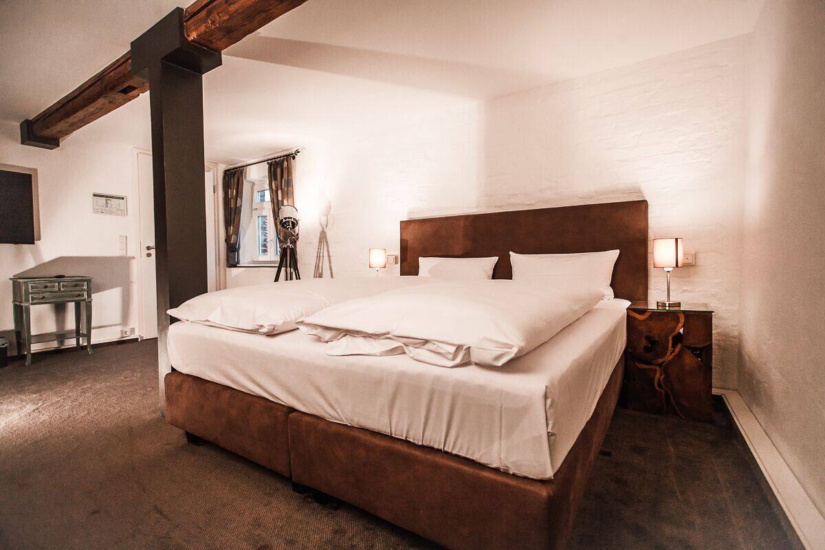 A neatly made double bed with a wooden headboard, a bedside table with a lamp, and a view of a window with curtains.