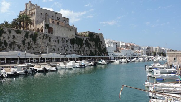 Un port de plaisance sur la côte, avec des bateaux amarrés, un bâtiment historique doté d’un mur fortifié et un ciel dégagé.