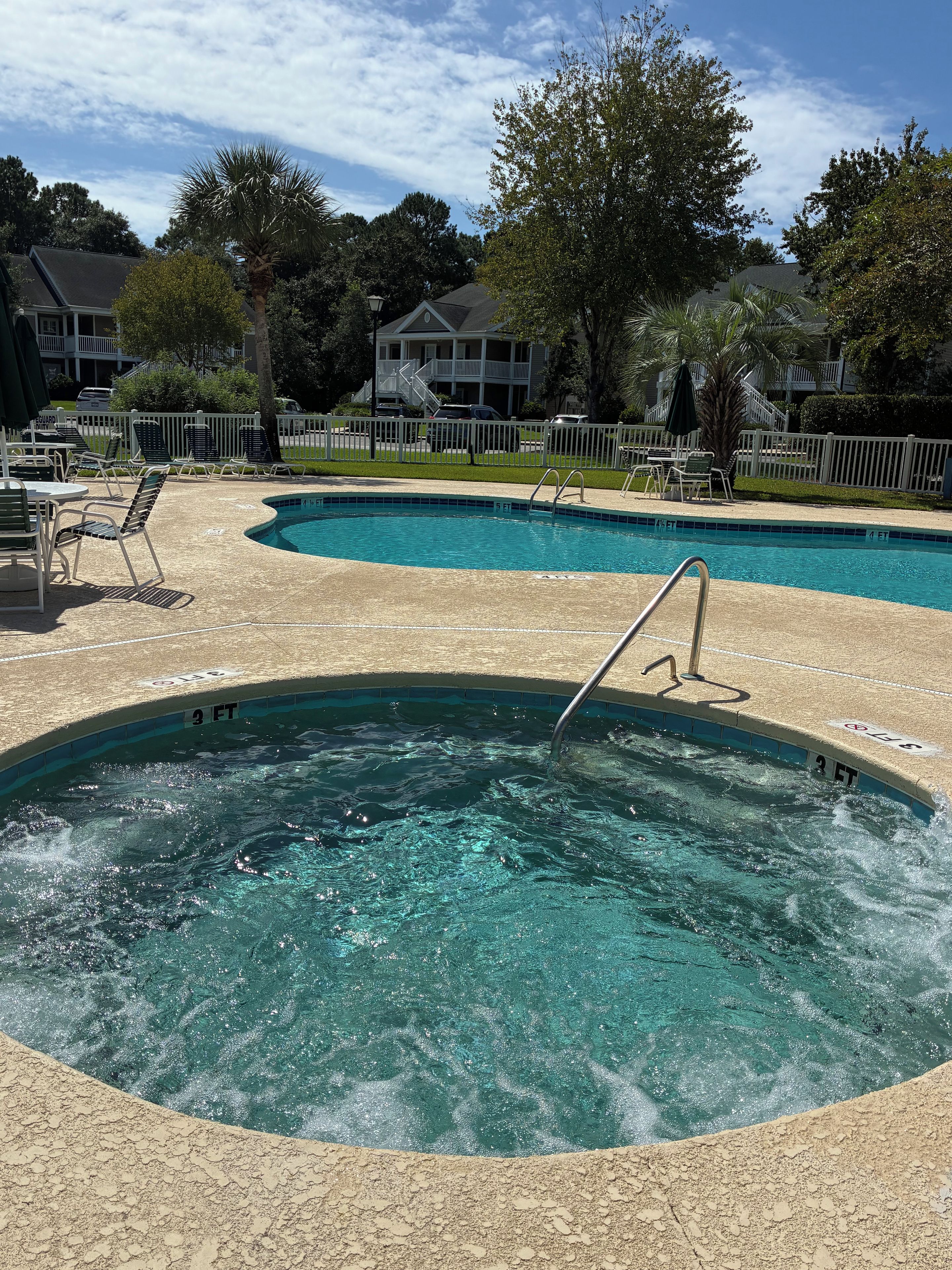 A swimming pool with a hot tub, surrounded by lounge chairs and a white fence.