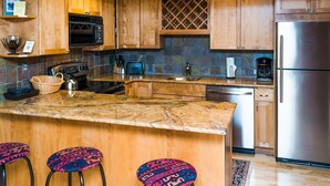 A modern kitchen with wooden cabinets, a stainless steel refrigerator, a microwave, and a coffee maker. There are three bar stools with colorful seats. - Lift One - Stylish three bedroom with walk-out patio (Aspen)
