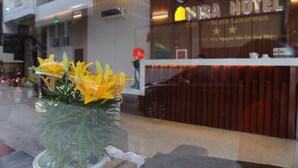 A hotel reception desk with a sign that reads "NABA HOTEL" and a clock above it. There is a potted plant with yellow flowers in the foreground. - Mira Hotel (Quy Nhon Nam)