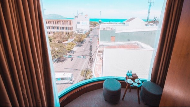 A room with a curved window overlooking a street and buildings, a small table with chairs, and a view of the ocean. - Mira Hotel (Quy Nhon Nam)