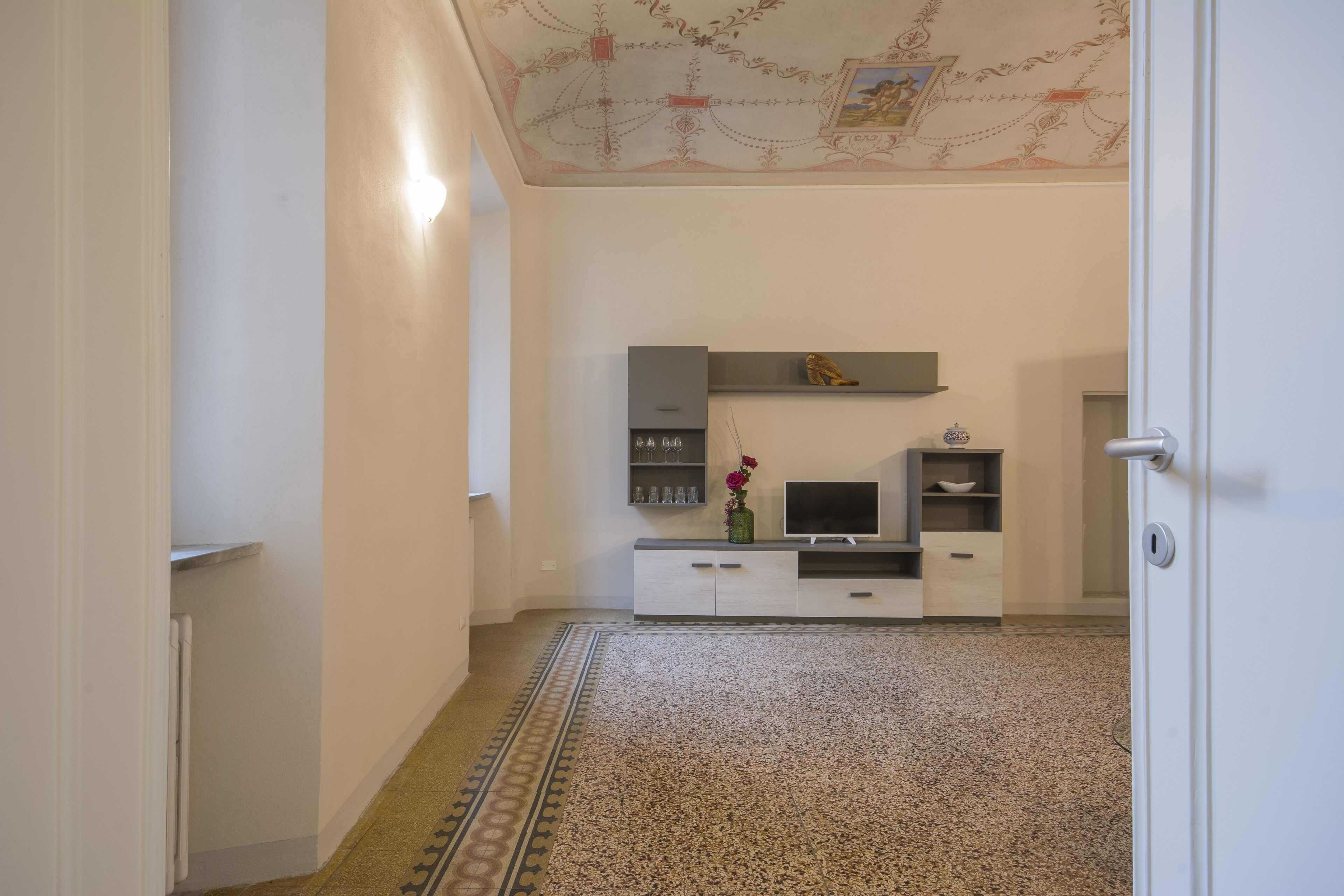 Un couloir avec un tapis à motifs, une porte blanche et une étagère fixée au mur, ornée d’objets décoratifs.