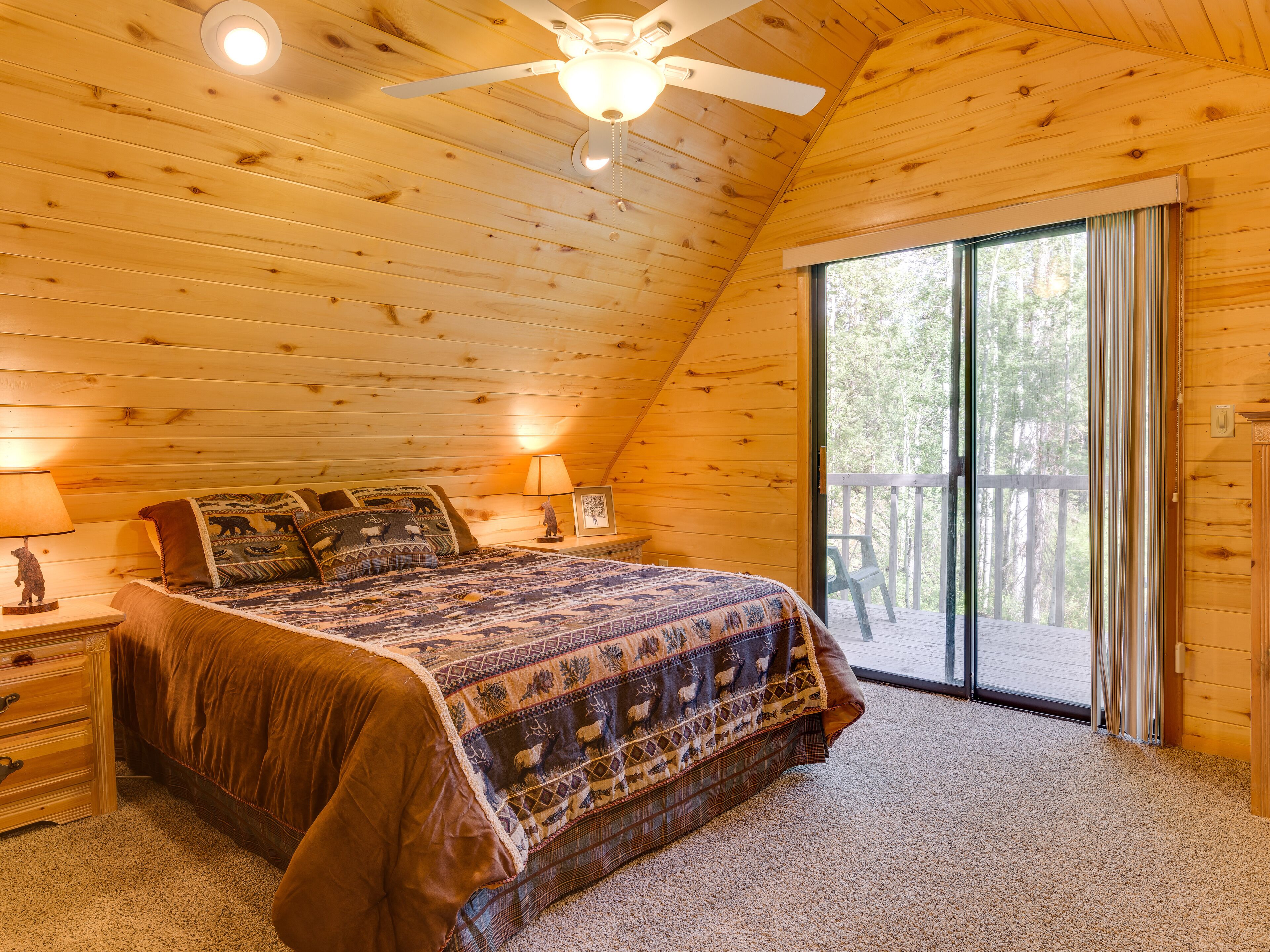 A cozy bedroom with a wooden ceiling, a large bed with patterned bedding, a nightstand with a lamp, and a sliding glass door leading to a balcony.