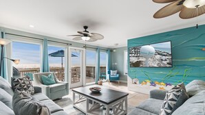 A spacious living room with a large gray sofa, a coffee table, and a ceiling fan. There is a large window with a view of the beach and a mural of the ocean on the wall. - Breezeway-Gulf Front Home Sleeps 9 (Navarre)