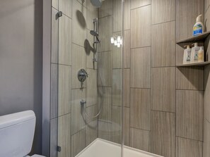 Doccia con porta in vetro, soffione a mano e rubinetto montato sulla parete. Si vede uno scaffale con articoli da toilette.