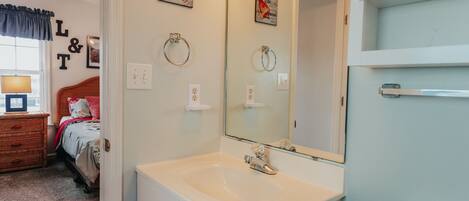 A bathroom with a white sink, mirror, and toilet. There is a bed with a headboard and a window with a curtain.