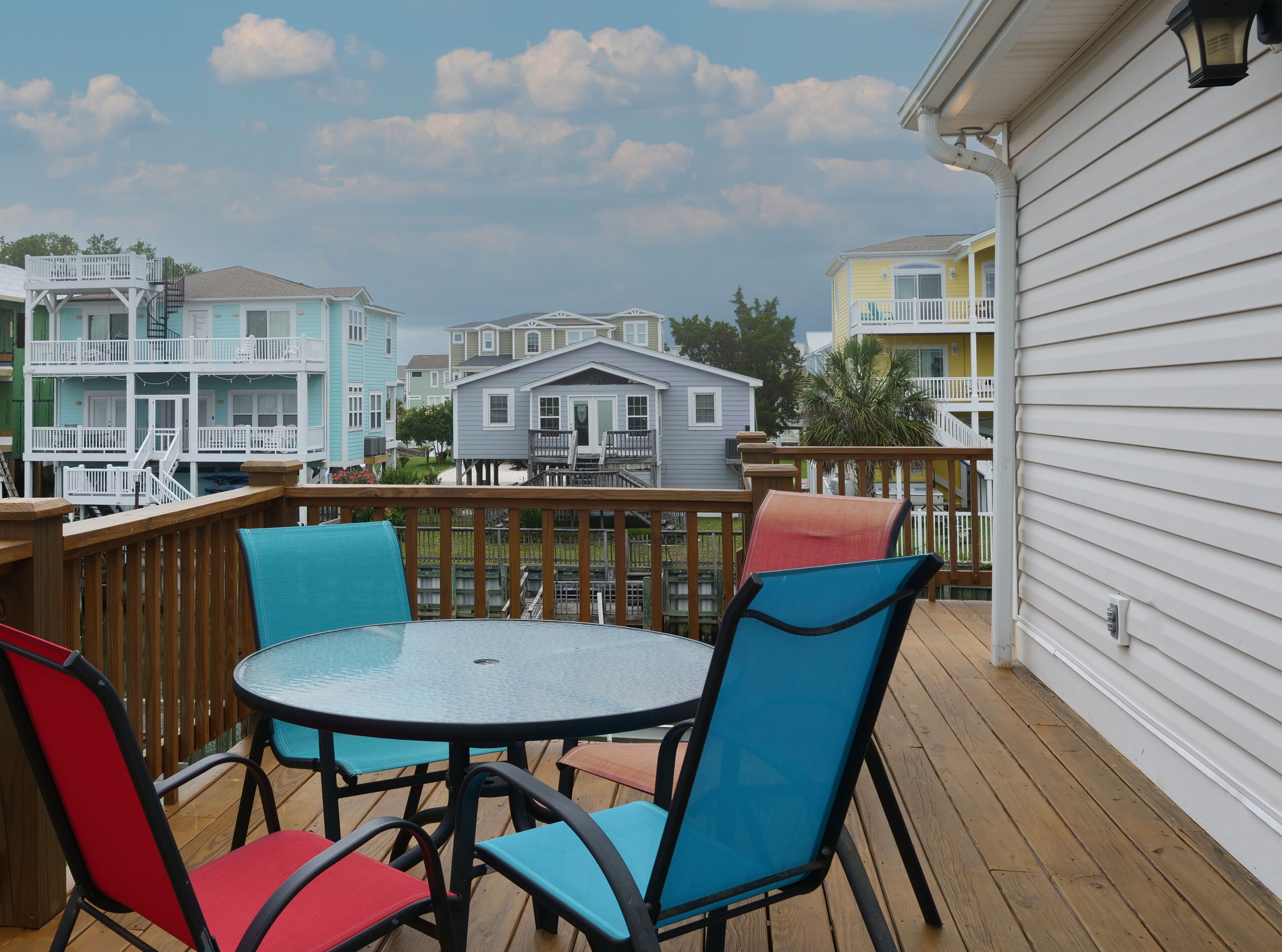 A wooden deck with a round table and colorful chairs, overlooking a row of houses with balconies.