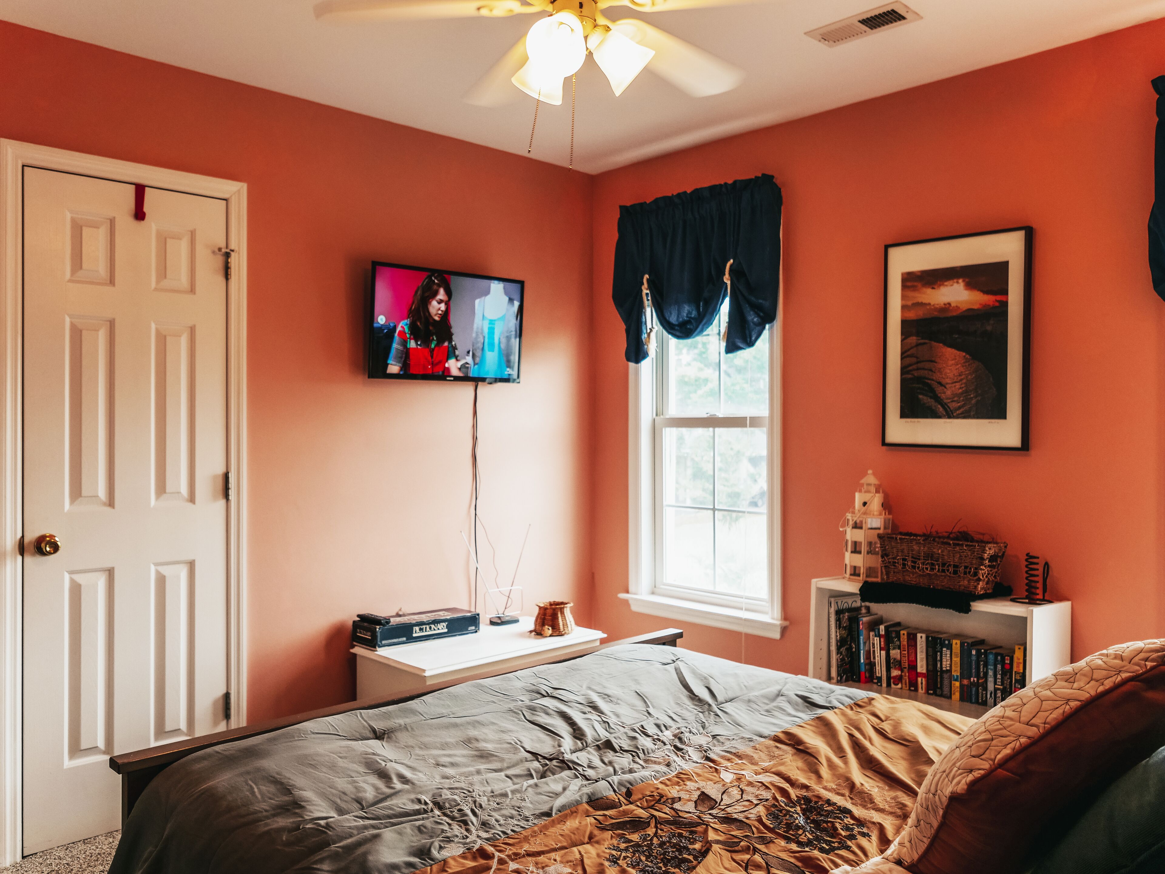 Ein Schlafzimmer mit einem Bett, einem an der Wand montierten Fernseher, einem Fenster mit Vorhang, einem Bücherregal und einer Tür.