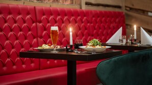 A red upholstered booth with a table set for two, featuring a glass of beer, a candle, and plates of food. - Bodetaler Basecamp Lodge (Oberharz am Brocken)