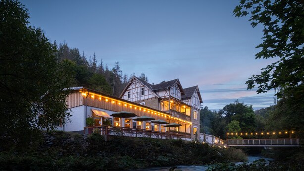 A traditional half-timbered building with outdoor seating and a river in the foreground. - Bodetaler Basecamp Lodge (Oberharz am Brocken)