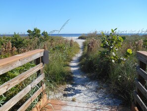Un sentier sablonneux menant à une plage, bordé de rambardes en bois et entouré de végétation.