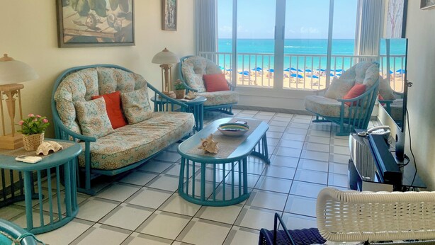A spacious living room with a view of the beach, featuring two patterned armchairs, a coffee table, and a television. - DREAM ESCAPE - TWO BEDROOM OCEANFRONT 
SEAS THE DAY, THE OCEAN IS YOUR NEIGHBOR. (Isla Verde)