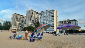 A beach with multiple high-rise buildings in the background, beach chairs, umbrellas, and a cooler. - DREAM ESCAPE - TWO BEDROOM OCEANFRONT 
SEAS THE DAY, THE OCEAN IS YOUR NEIGHBOR. (Isla Verde)