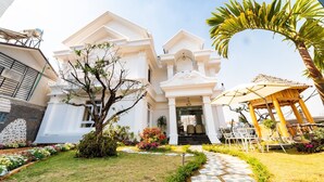 A white building with a symmetrical facade, a central entrance with steps, and a covered outdoor seating area. - King Villa (Xuan Huong - Da Lat)