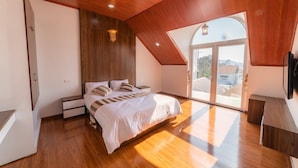 A modern bedroom with a wooden ceiling, a large bed, a nightstand, a TV, and a sliding glass door leading to a balcony. - King Villa (Xuan Huong - Da Lat)