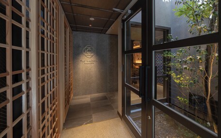 A corridor with a wooden lattice wall, a sign with "The Mashiya Kazahaya" logo, and a view of a brick building and trees through glass doors.