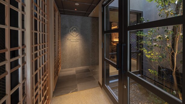 A corridor with a wooden lattice wall, a sign with "The Mashiya Kazahaya" logo, and a view of a brick building and trees through glass doors.