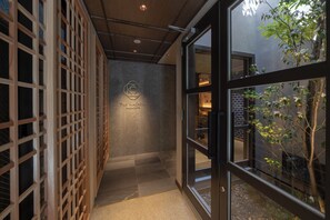 Un couloir avec un mur en treillis de bois, une enseigne portant le logo « The Mashiya Kazahaya », et une vue sur un bâtiment en briques et des arbres, visibles à travers des portes vitrées.