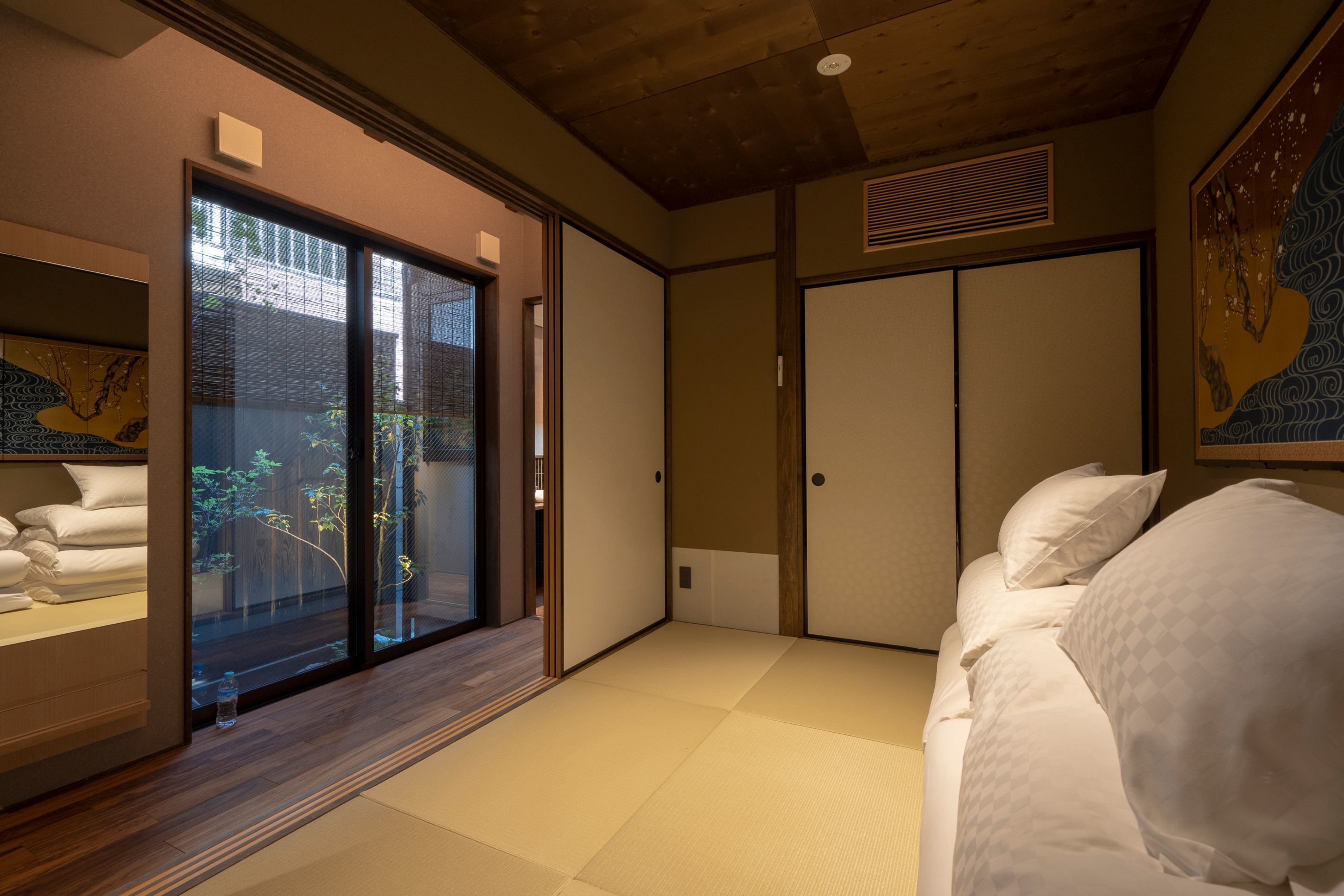 Ett hotellrum med tatamimattor på golvet, en säng med vita sängkläder, en skjutdörr som leder ut till en balkong och ett traditionellt japanskt konstverk på väggen.
