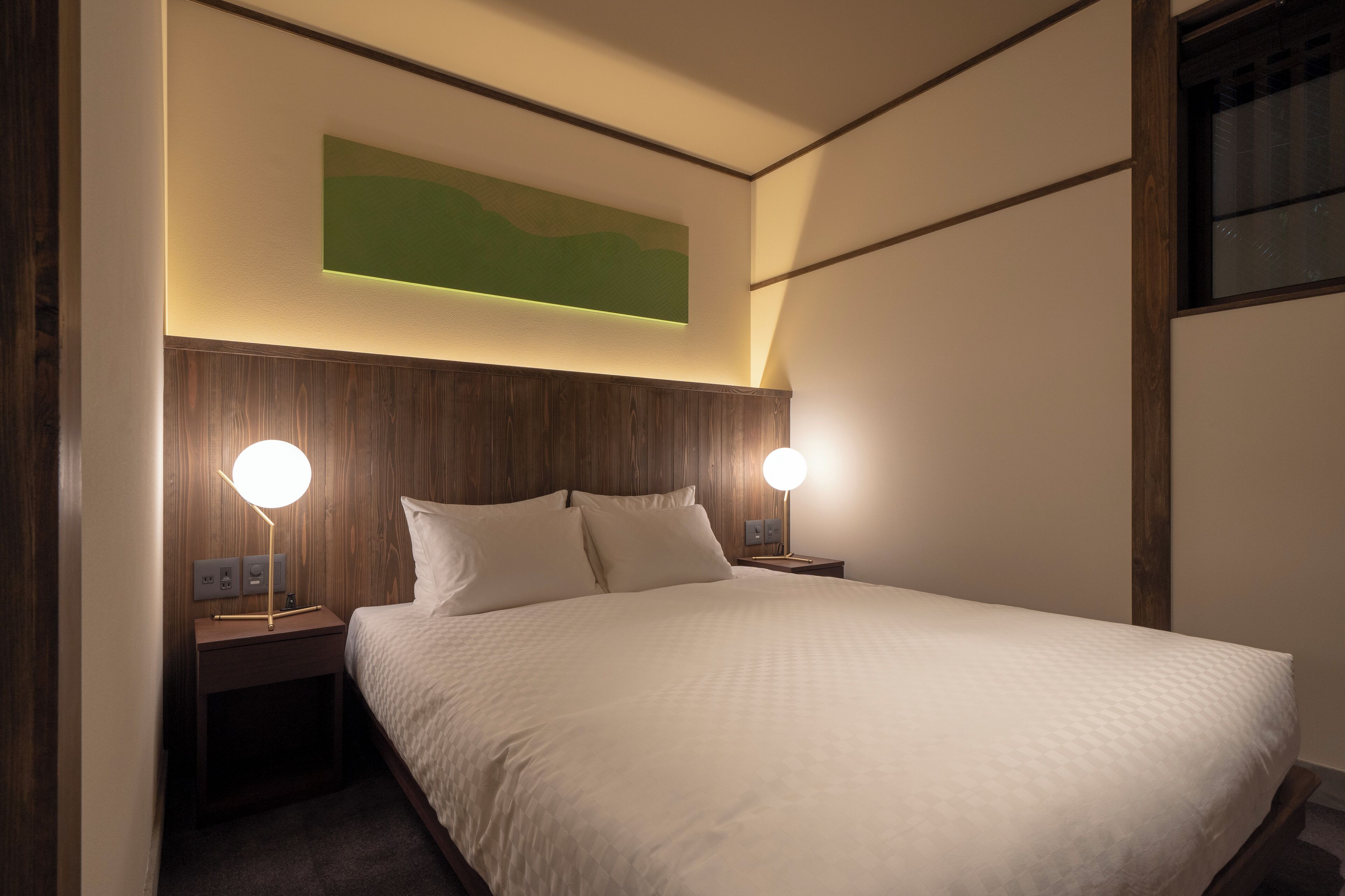 A neatly made bed with a white comforter and pillows, two bedside lamps, a wooden headboard, and a green abstract painting above the bed.