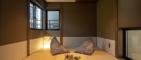 A traditional Japanese room with tatami flooring, two tatami mats, a small table with a teapot and cups, and a window with a view of a wooden structure outside.