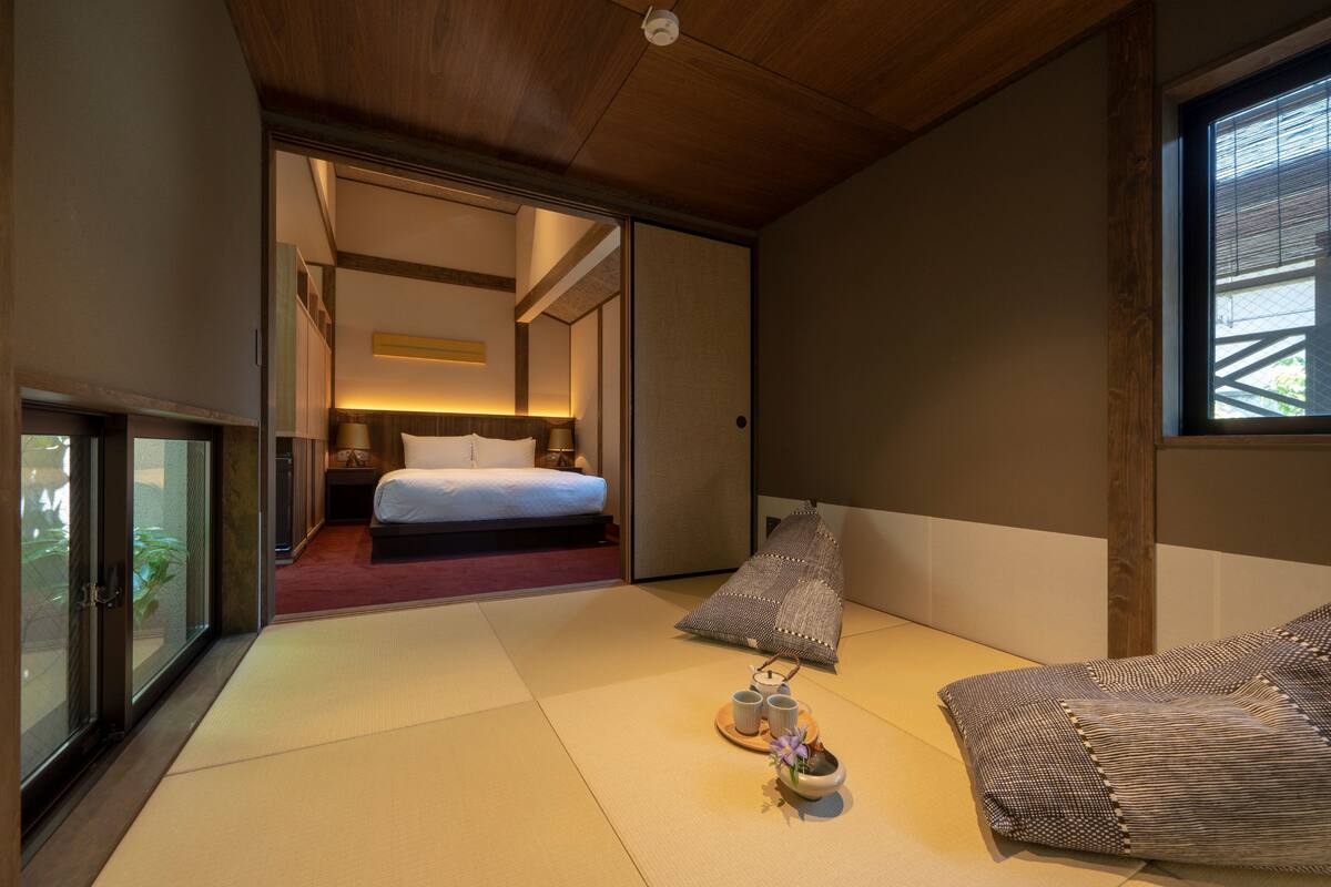 Ein traditioneller japanischer Raum mit Tatami-Böden, einem Bett mit weißer Matratze, einem Kissen und einem kleinen Tisch mit Tassen und einer Blume.