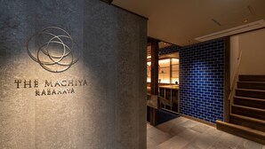A modern interior with a concrete wall displaying "THE MACHIYA KAZAHAYA" logo, a wooden staircase, and a blue-tiled wall.