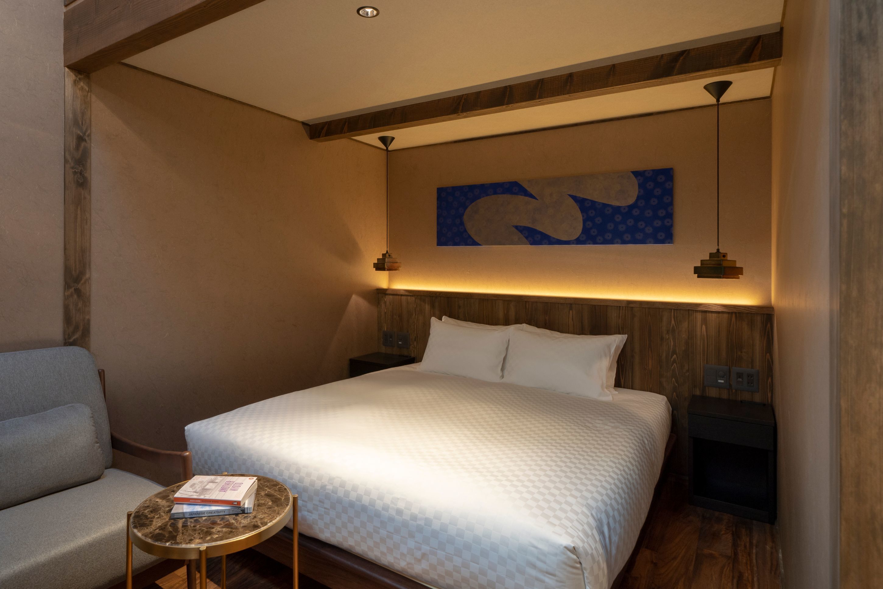 A hotel room with a bed, a small table with books, a nightstand, and a wall art piece.
