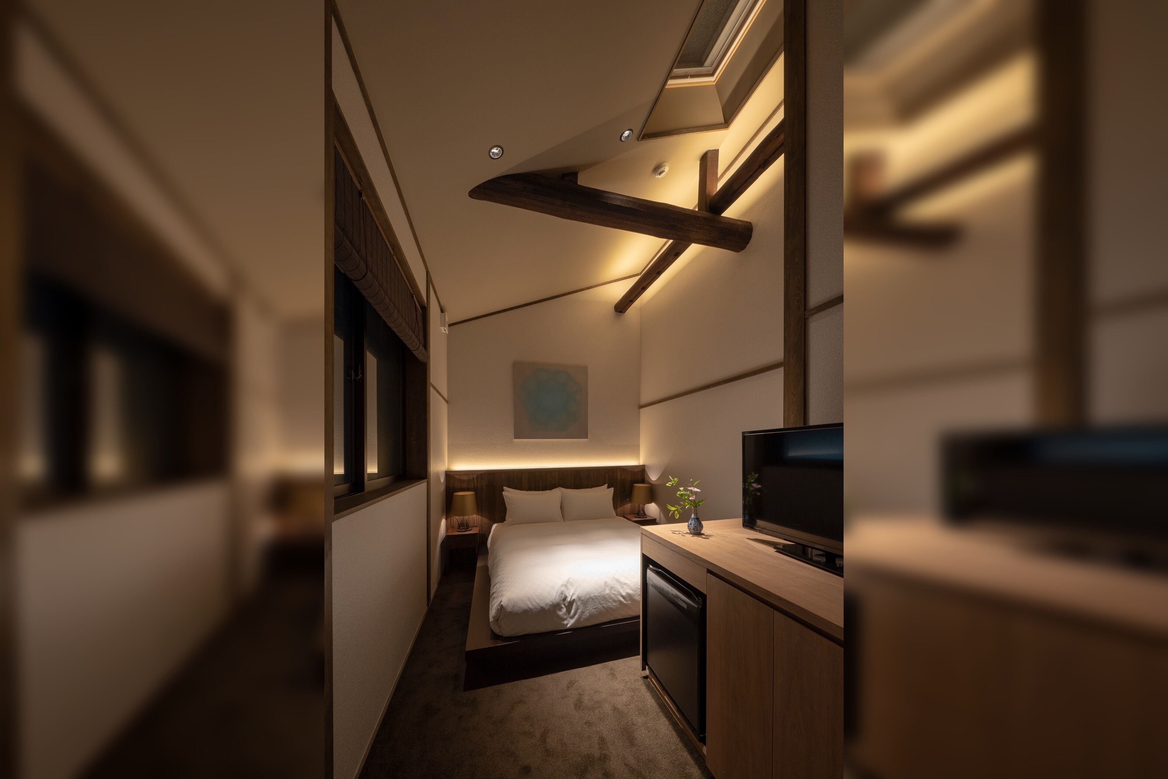 A narrow hotel room with a bed, bedside tables, a TV, and a small desk area.