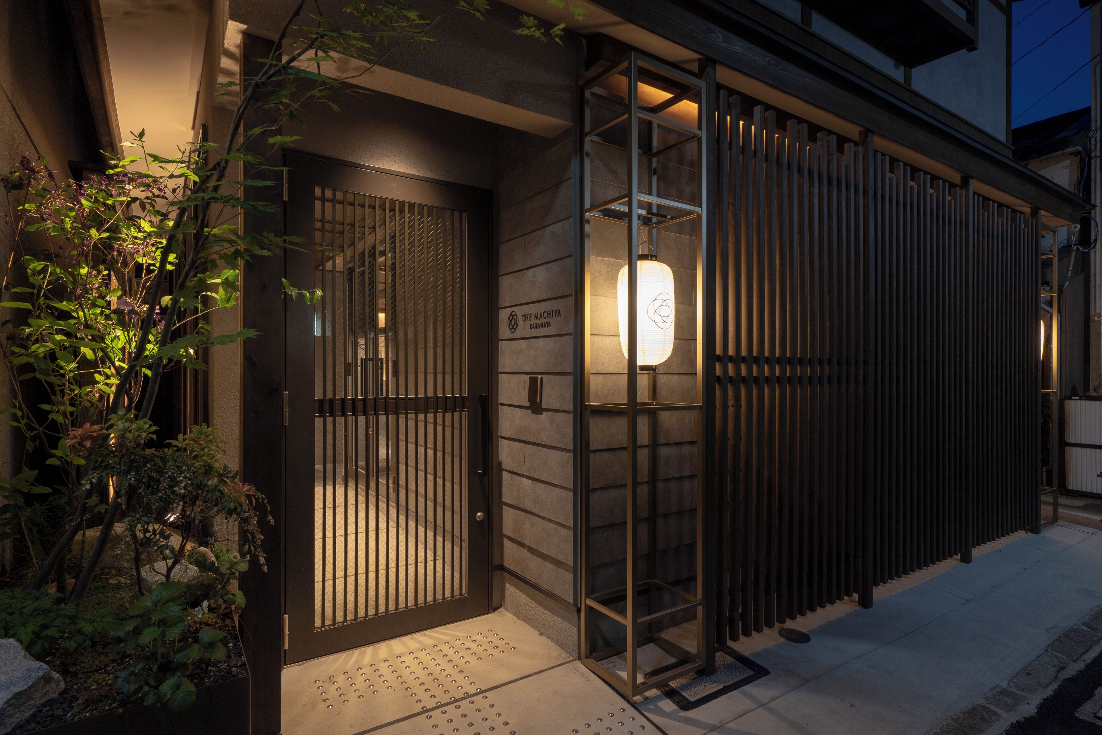 A modern building with a gated entrance, a lit lantern, and a sign reading "The Machiya".
