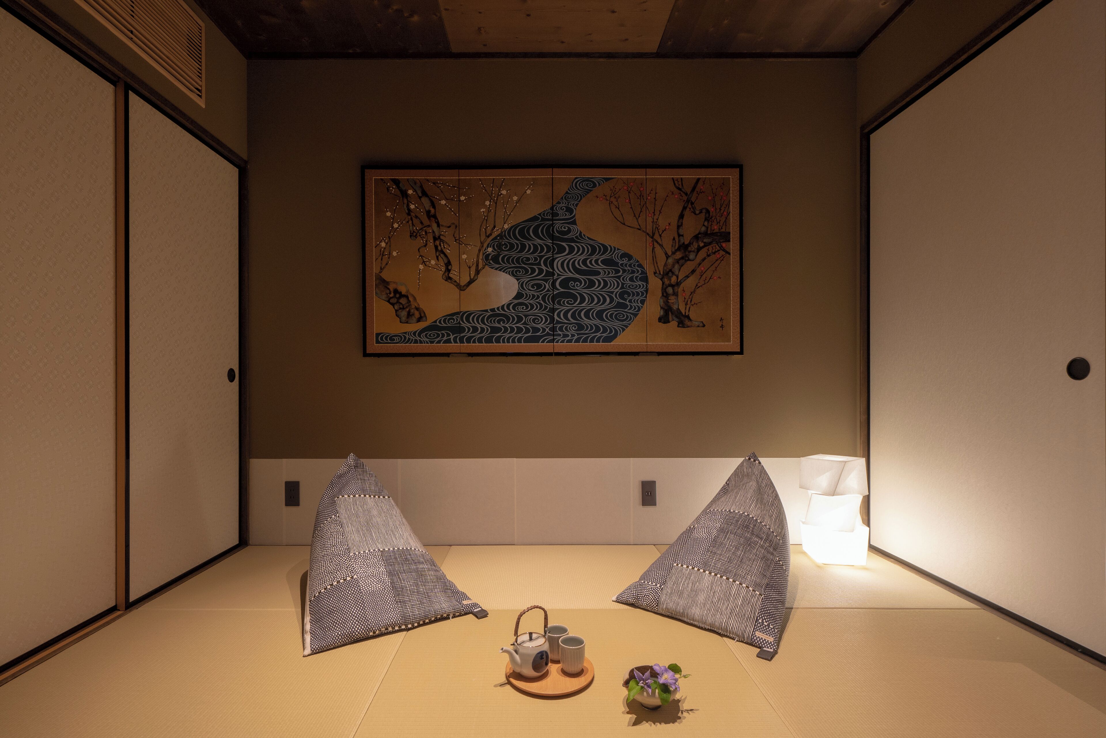 Une pièce avec un sol recouvert de tatamis, ornée d’une peinture japonaise traditionnelle, comprenant un service à thé et une petite plante.
