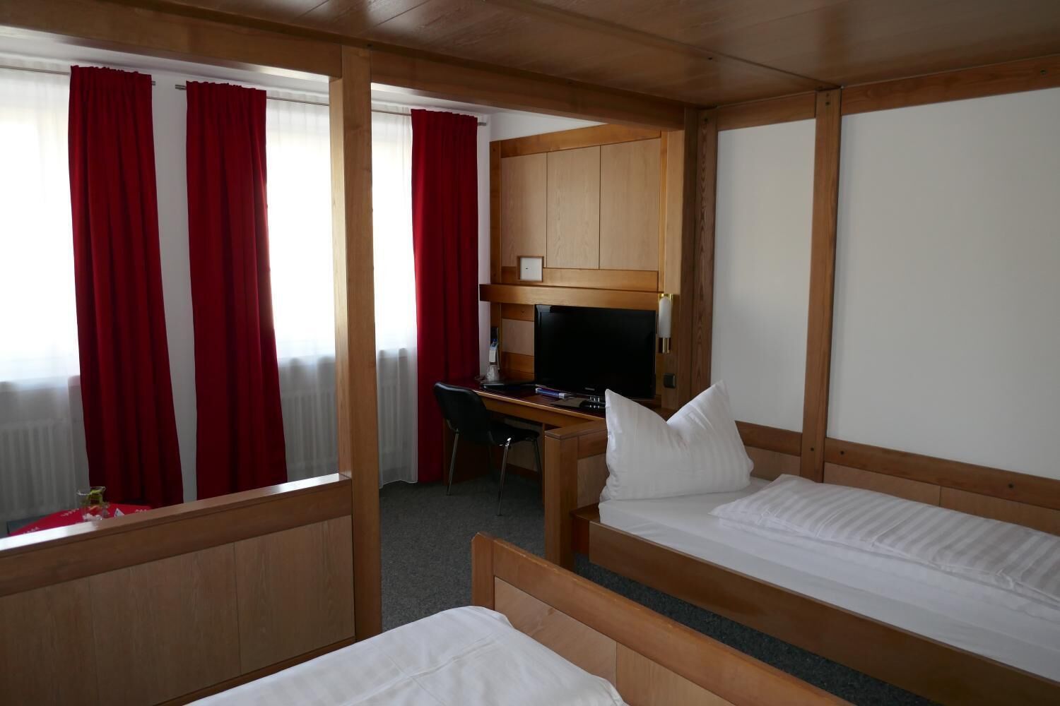 A hotel room with two single beds, a wooden desk with a television, and a window with red curtains.