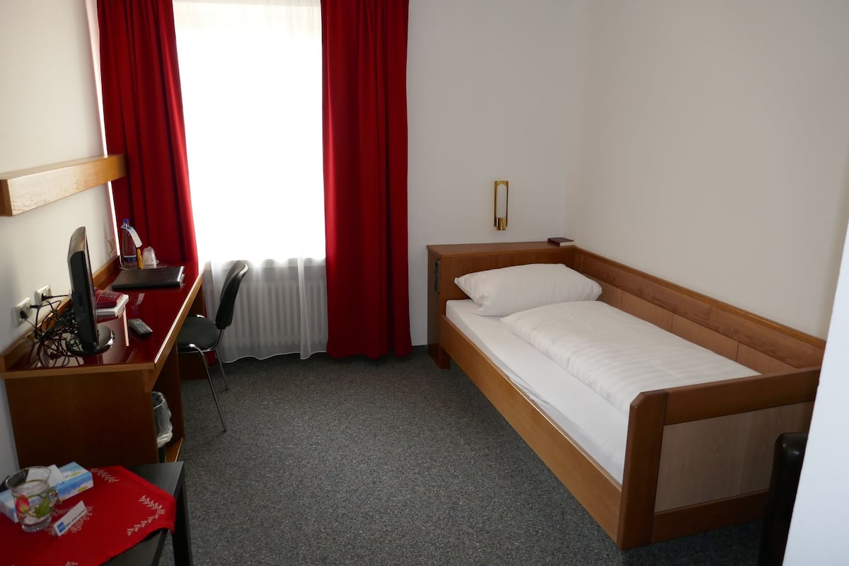Een enkel bed met houten frame en witte beddengoed, een bureau met stoel, een computer en een klein tafeltje met een rode tafelkleed.