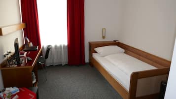 Una cama individual con estructura de madera y ropa de cama blanca, un escritorio con una silla, una computadora y una mesita con un mantel rojo.
