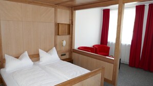 A hotel room with a wooden bed, a small bedside table, a mirror, and a window with red curtains. - Hotel Bayerischer Hof (Burghausen)