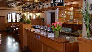 A bar area with a wooden counter, bottles of wine, and glasses. There are tables and chairs in the background. - Hotel Bayerischer Hof (Burghausen)