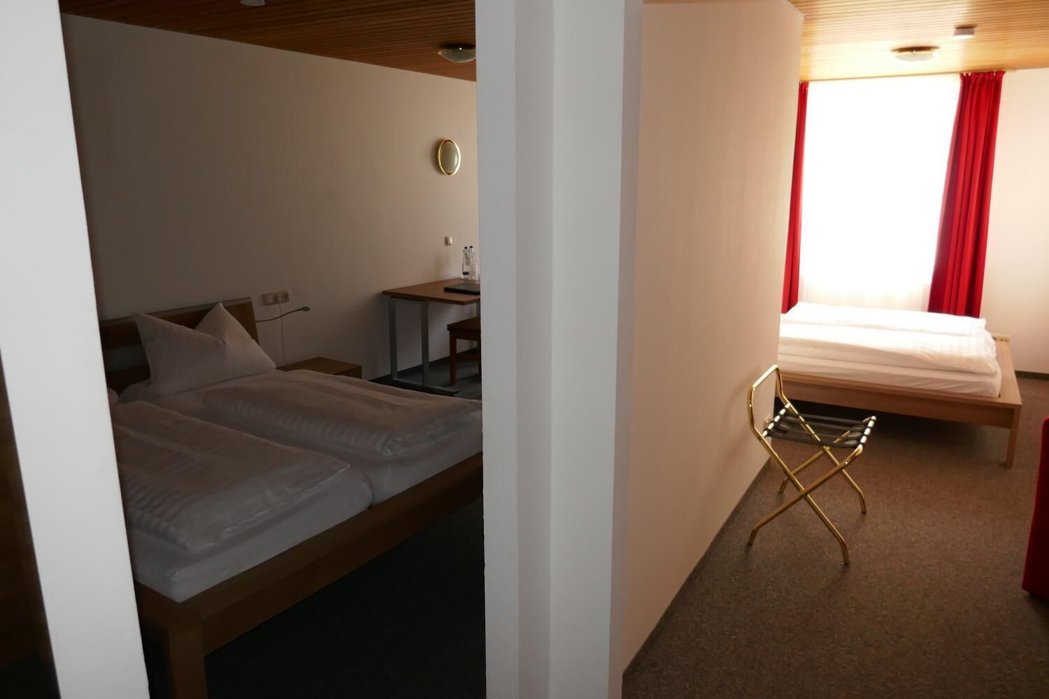 A hotel room with a double bed, a desk with a chair, a small table, and a window with red curtains.