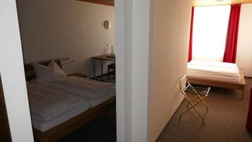 A hotel room with a double bed, a desk with a chair, a small table, and a window with red curtains.